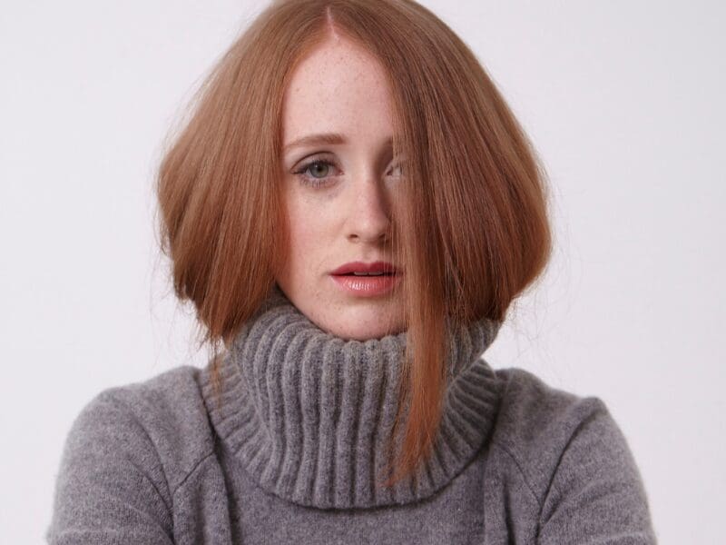 Woman with straight, shoulder-length copper-red hair falling over one eye, freckles and coral lips, in a gray turtleneck.