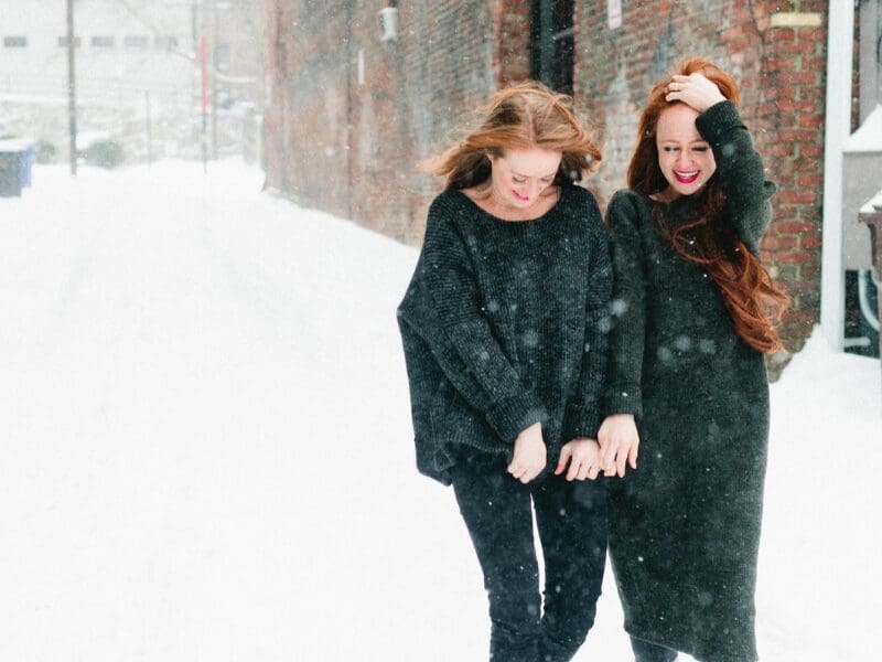 Two laughing redheads in dark knit sweaters walking arm in arm down a snowy alley beside a brick wall as snow falls.